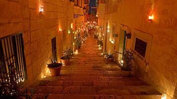 Une ruelle étroite, pavée de pierres, agrémentée de plantes en pot et éclairée d’une lumière chaleureuse, qui mène à l’entrée d’un immeuble.