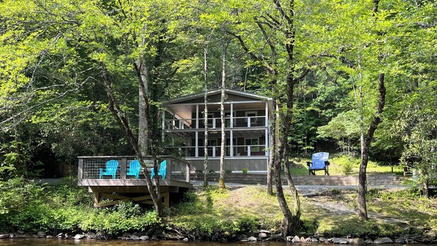 A two-story cabin with a deck overlooking a clear stream, surrounded by dense greenery. - Blue Ridge Riverfront Cabin•2-Level Deck•Sleeps 8 (Blue Ridge)