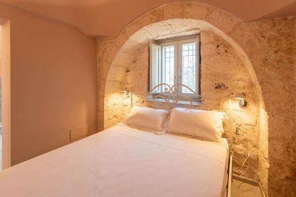 Une chambre avec un mur en pierre, une fenêtre, un lit avec du linge de lit blanc et des lampes fixées au mur.