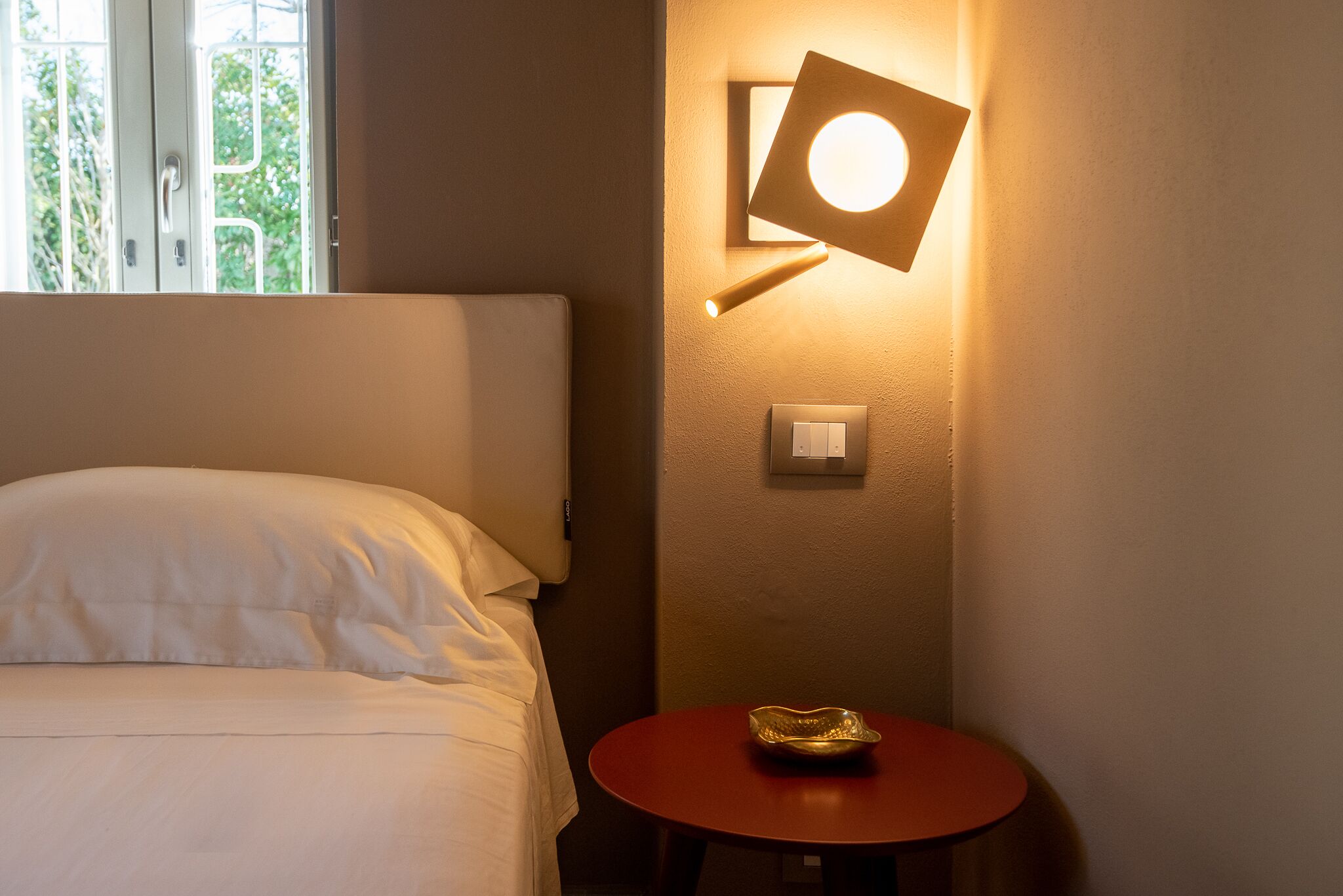 Habitación de hotel con cama, una mesa redonda con un bol, luz empotrada en la pared y ventana con vistas a la vegetación.