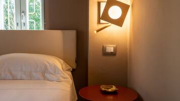 Habitación de hotel con cama, una mesa redonda con un bol, luz empotrada en la pared y ventana con vistas a la vegetación.