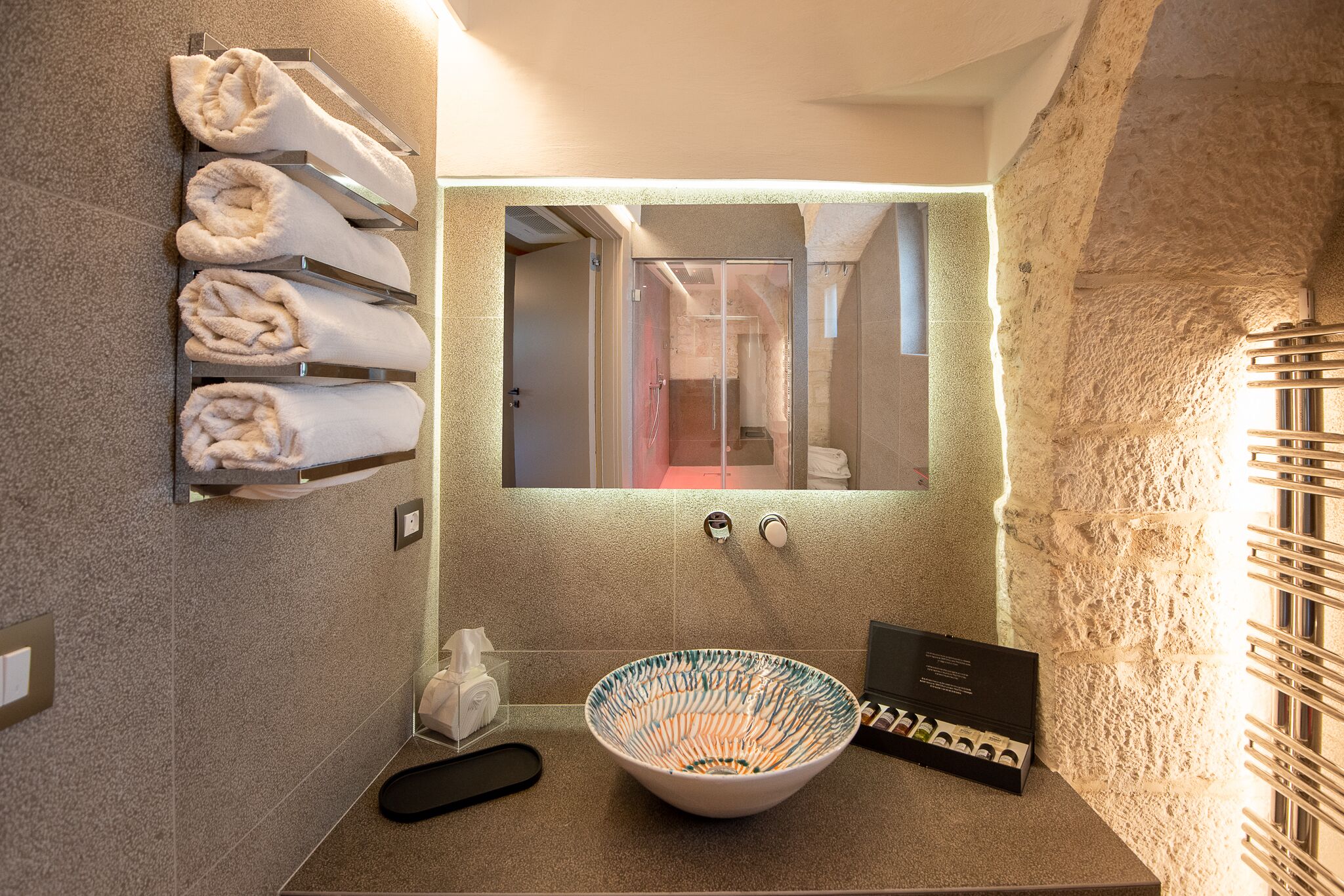 Un baño con pared de piedra, un espejo grande, un lavamanos con un cuenco estampado, un perchero con toallas dobladas y una botella de jabón para manos.