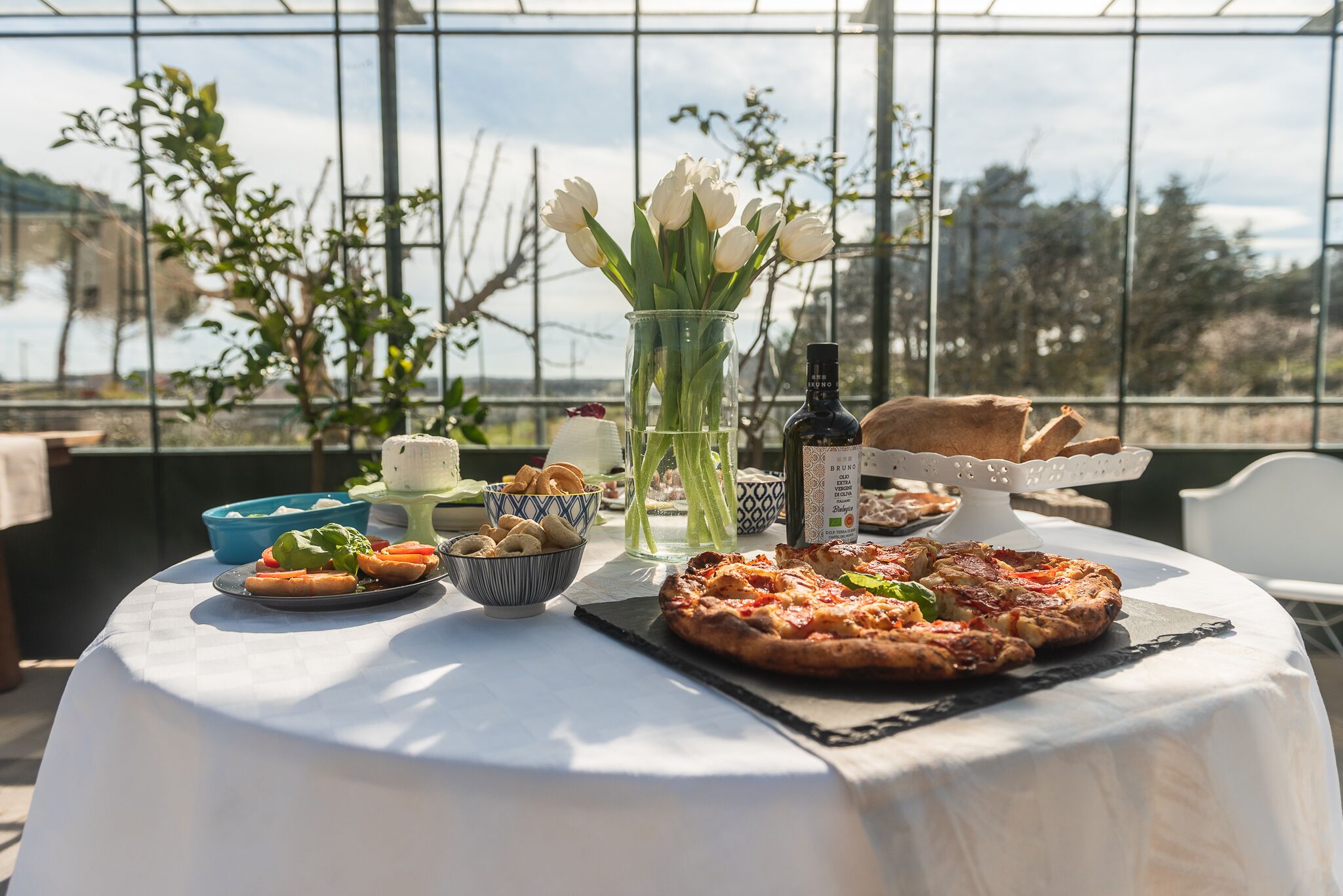 Una mesa con pizza, ensalada, pan y un jarrón de tulipanes en una terraza con vistas.