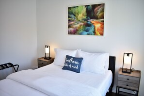A neatly made bed with white linens, a pillow with text, bedside lamps, and a colorful abstract painting on the wall. - The Worth Modern: Downtown 3BR home (Raleigh)