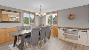 Un espace repas comprenant une table en bois, des chaises à revêtement gris, un lustre, de grandes fenêtres et une cheminée.