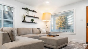 A living room with a sectional sofa, an ottoman, a floor lamp, and a large window with blinds.