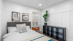 A bedroom with a bed, bedside table, lamp, and a black console unit against a white wall.