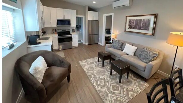 A modern living room with a brown armchair, a grey sofa, and a patterned rug. A kitchen with white cabinets and stainless steel appliances is visible in the background. - Chic & intimate 2 bedroom (Brighton)
