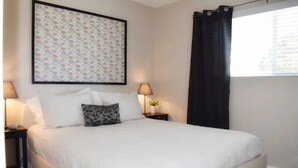 Una cama bien tendida con ropa blanca y un cojín decorativo estampado, una obra de arte enmarcada encima de la cama y una ventana con cortinas negras.