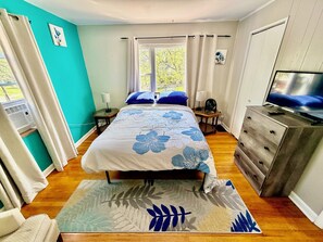 A bedroom with a bed, a TV, a dresser, a window, and a fan.