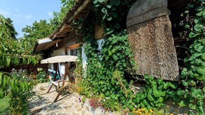 A rustic stone pathway leading to a wooden house with a thatched roof, surrounded by lush greenery and flowering plants.