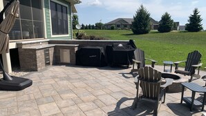 Une terrasse extérieure avec un sol en pierre, un barbecue, des fauteuils Adirondack et un foyer.