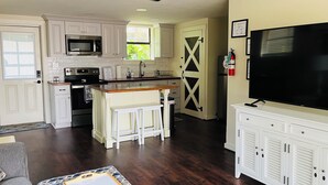A modern kitchen with white cabinets, a microwave, stove, and a large TV on a cabinet. There is a wooden dining table and a bar stool. - Maplewood Haven (Daphne)