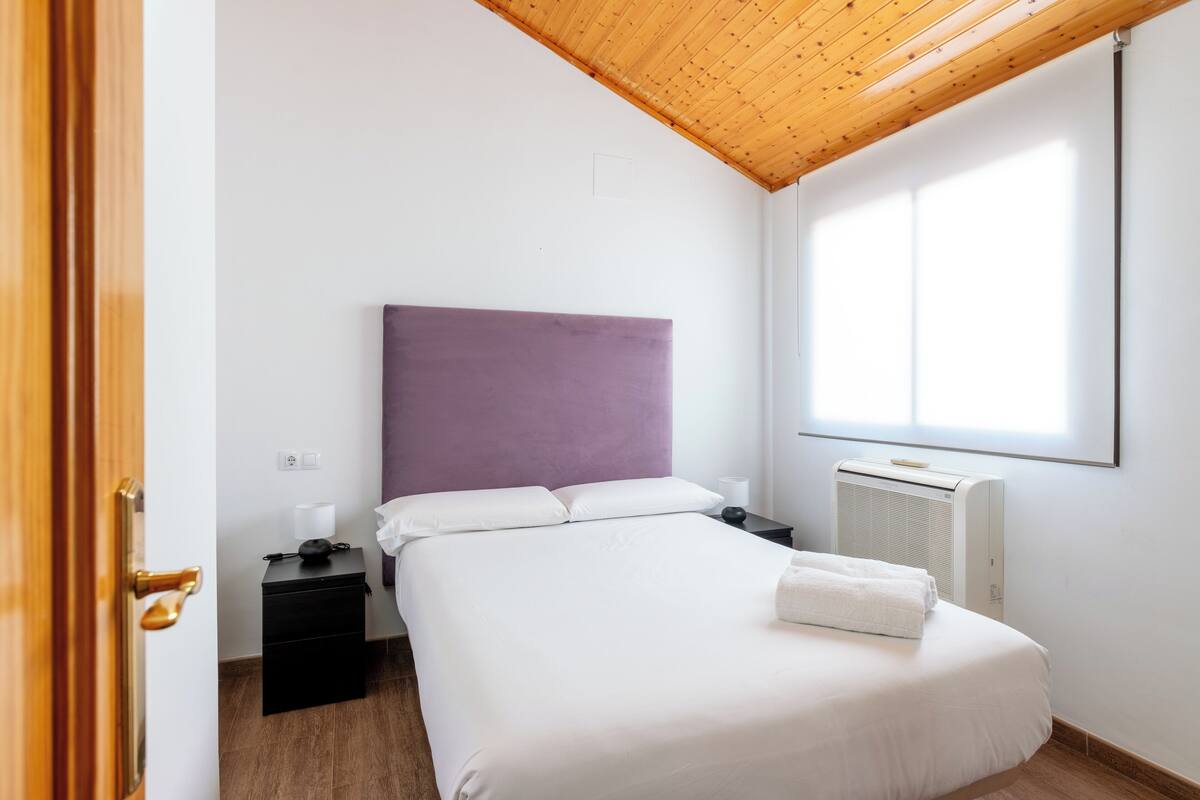 A bedroom with a wooden ceiling, a bed with white bedding, a purple headboard, a bedside table with a lamp, and a window with blinds.