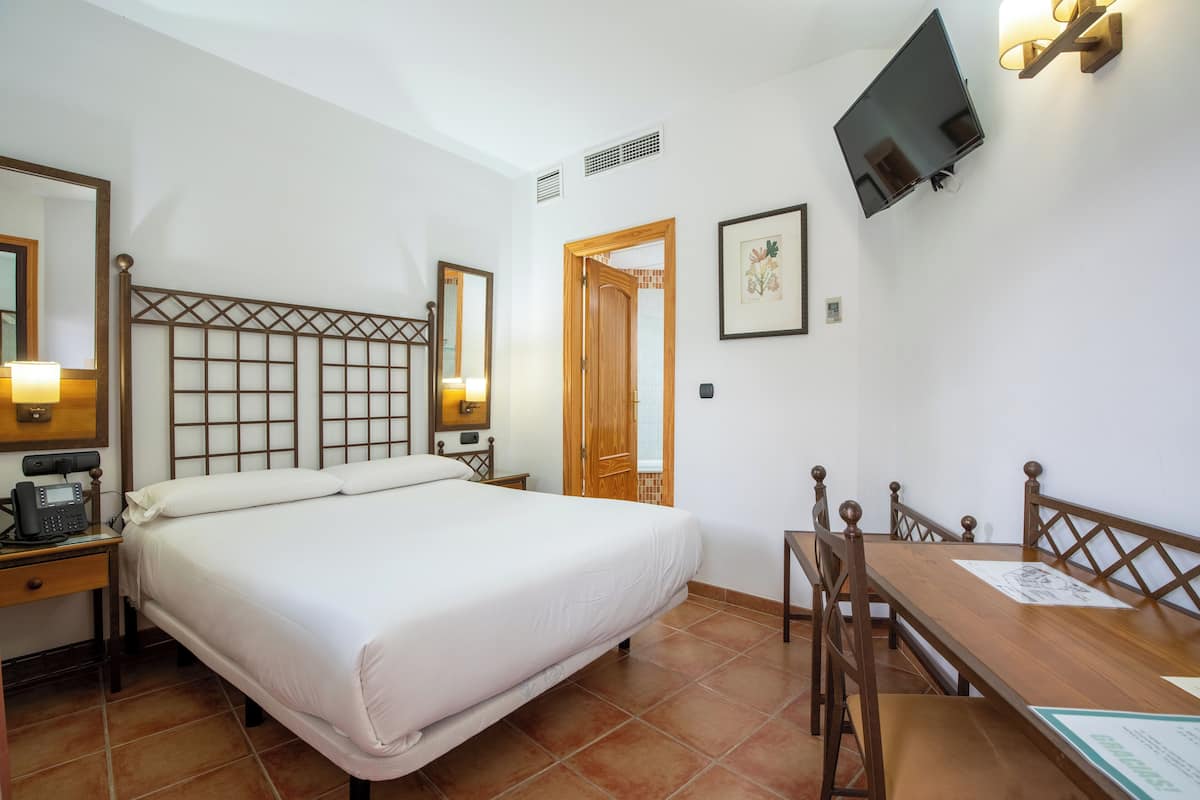 A hotel room with a bed, bedside table with a phone, a wooden desk with chairs, a TV mounted on the wall, and a door leading to another room.