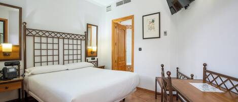 A hotel room with a bed, bedside table with a phone, a wooden desk with chairs, a TV mounted on the wall, and a door leading to another room.