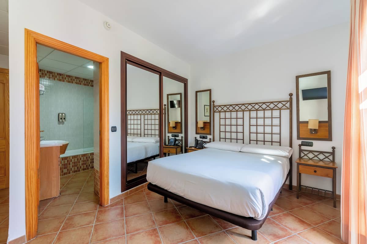 A bedroom with a large bed, a wooden headboard, a nightstand, a mirror, and a bathroom visible through an open door.