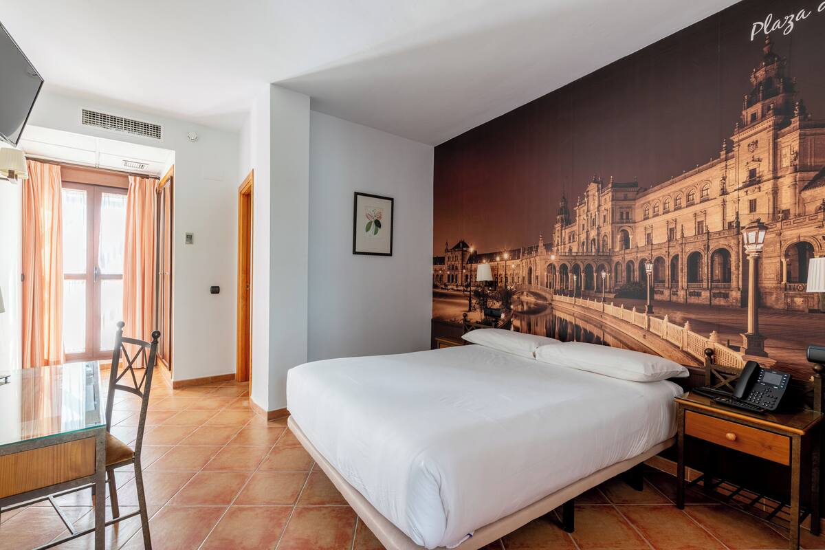 A hotel room with a large bed, a desk with a chair, a telephone, and a wall mural of a building.