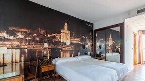 A hotel room with a bed, a desk, and a chair. There is a large wall mural of a cityscape.