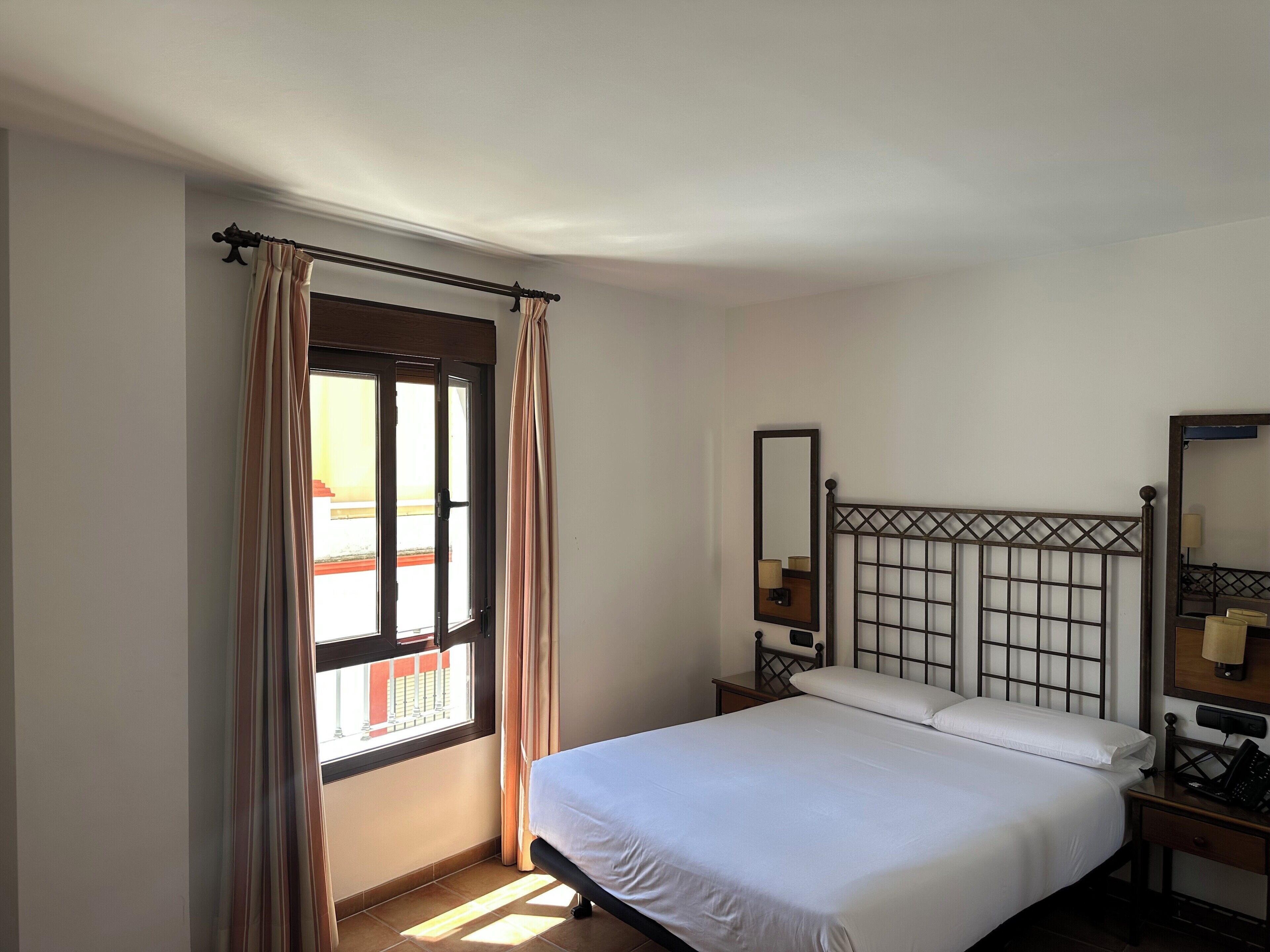 A bedroom with a bed, a window with curtains, a bedside table with a lamp, and a mirror on the wall.