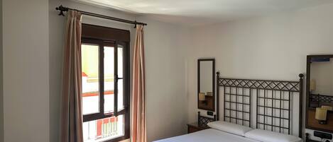 A bedroom with a bed, a window with curtains, a bedside table with a lamp, and a mirror on the wall.