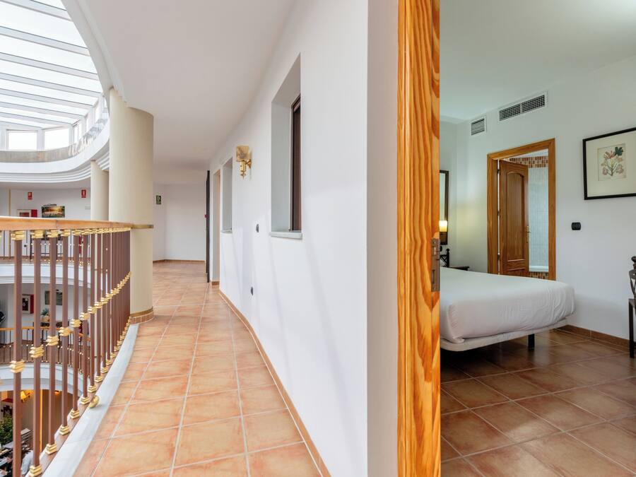 Un pasillo de hotel con techo alto, barandal de madera, una cama y una puerta de madera.