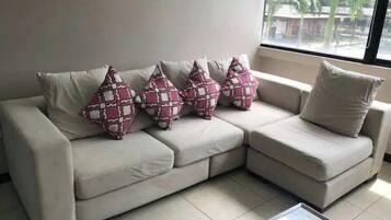 A beige sectional sofa with four patterned throw pillows and a plain pillow, positioned near a window with a view of palm trees.