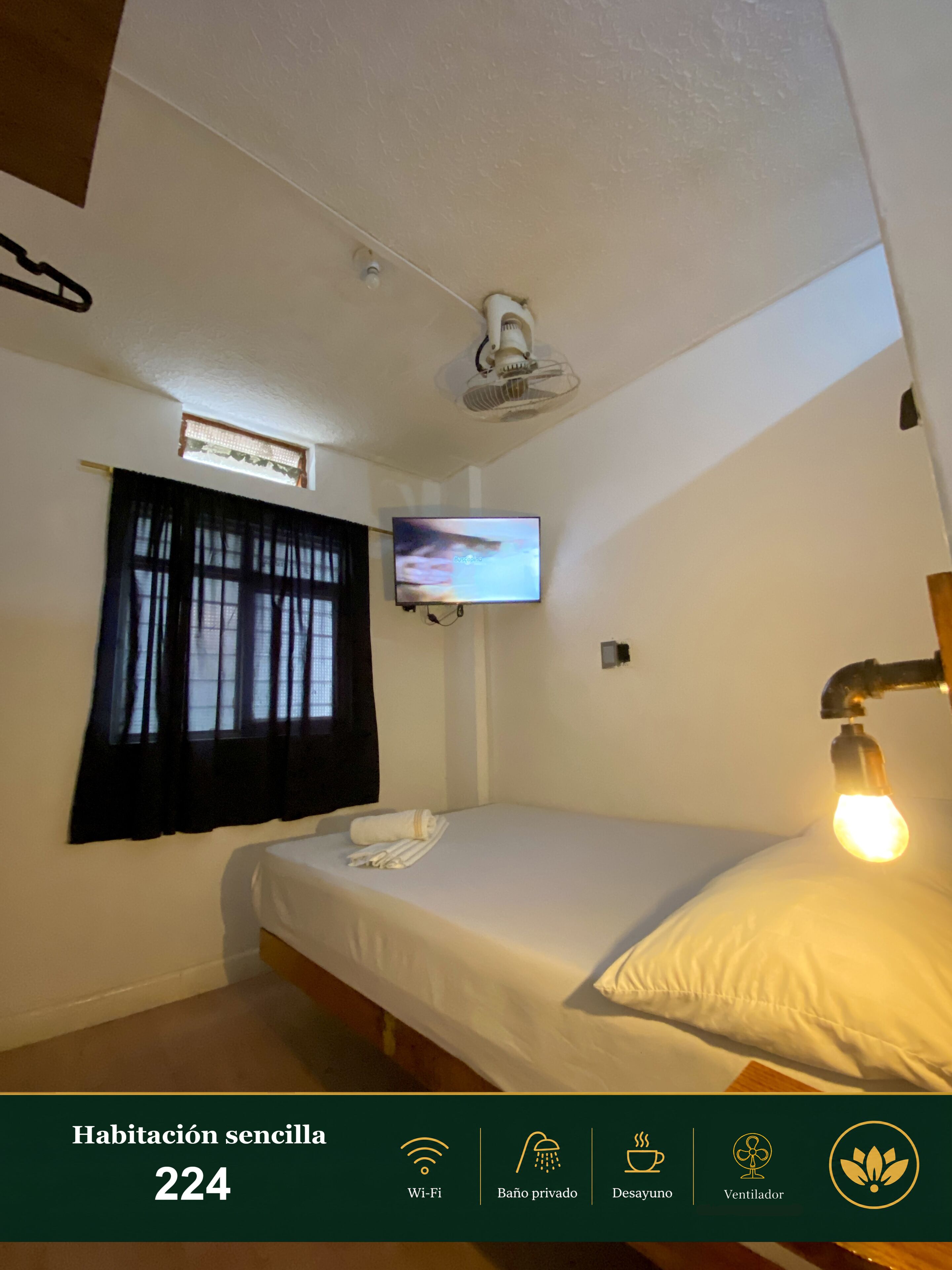 Une pièce simple, avec un lit, une télévision, un ventilateur de plafond et une fenêtre avec des rideaux.