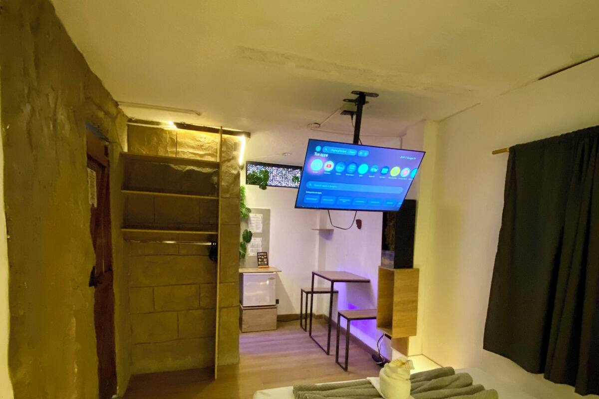 A hotel room with a bed, a TV mounted on the wall, a desk, and a small kitchenette.