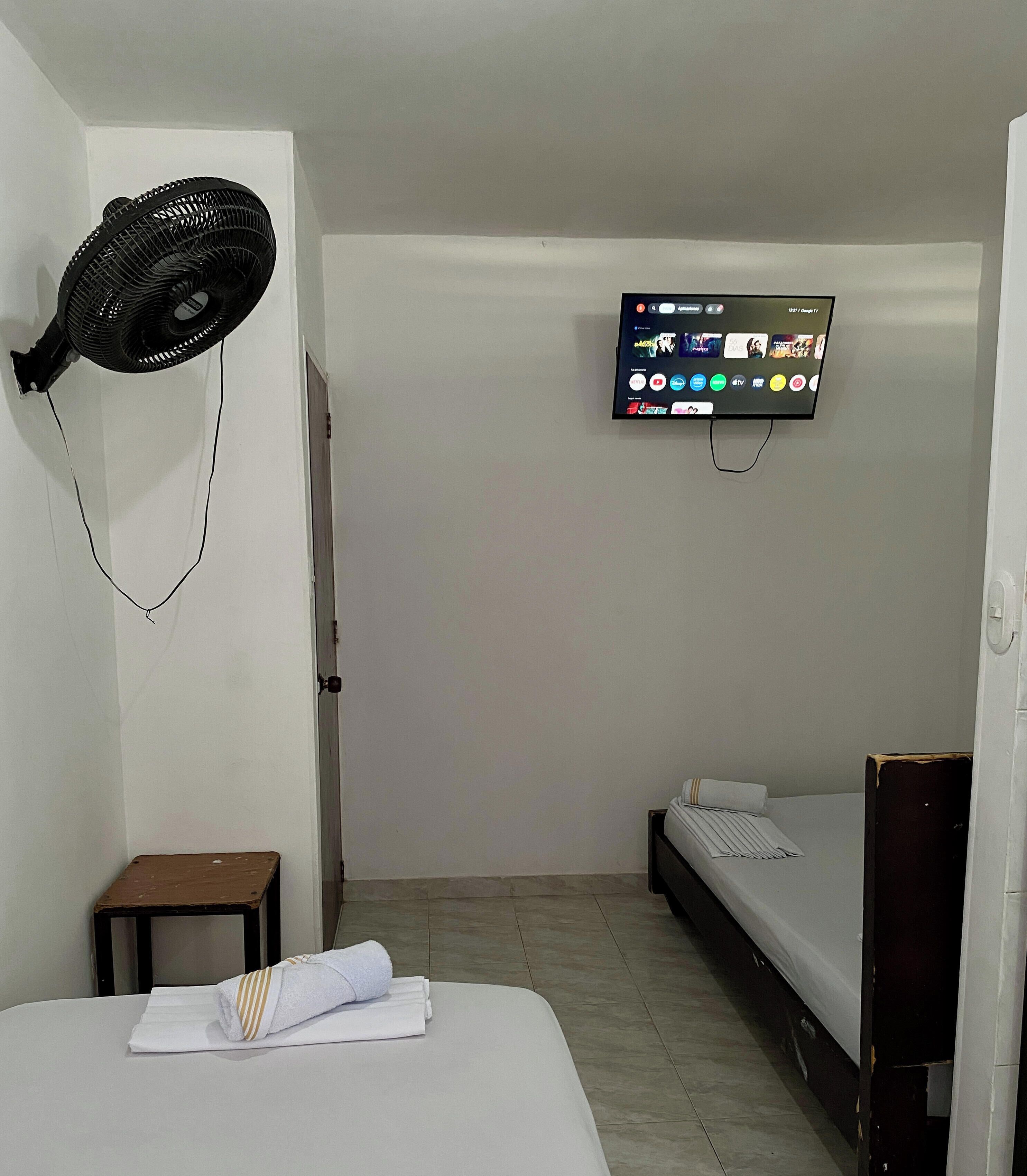 A small, clean room with a bed, a bedside table, a wall-mounted TV, and a standing fan.