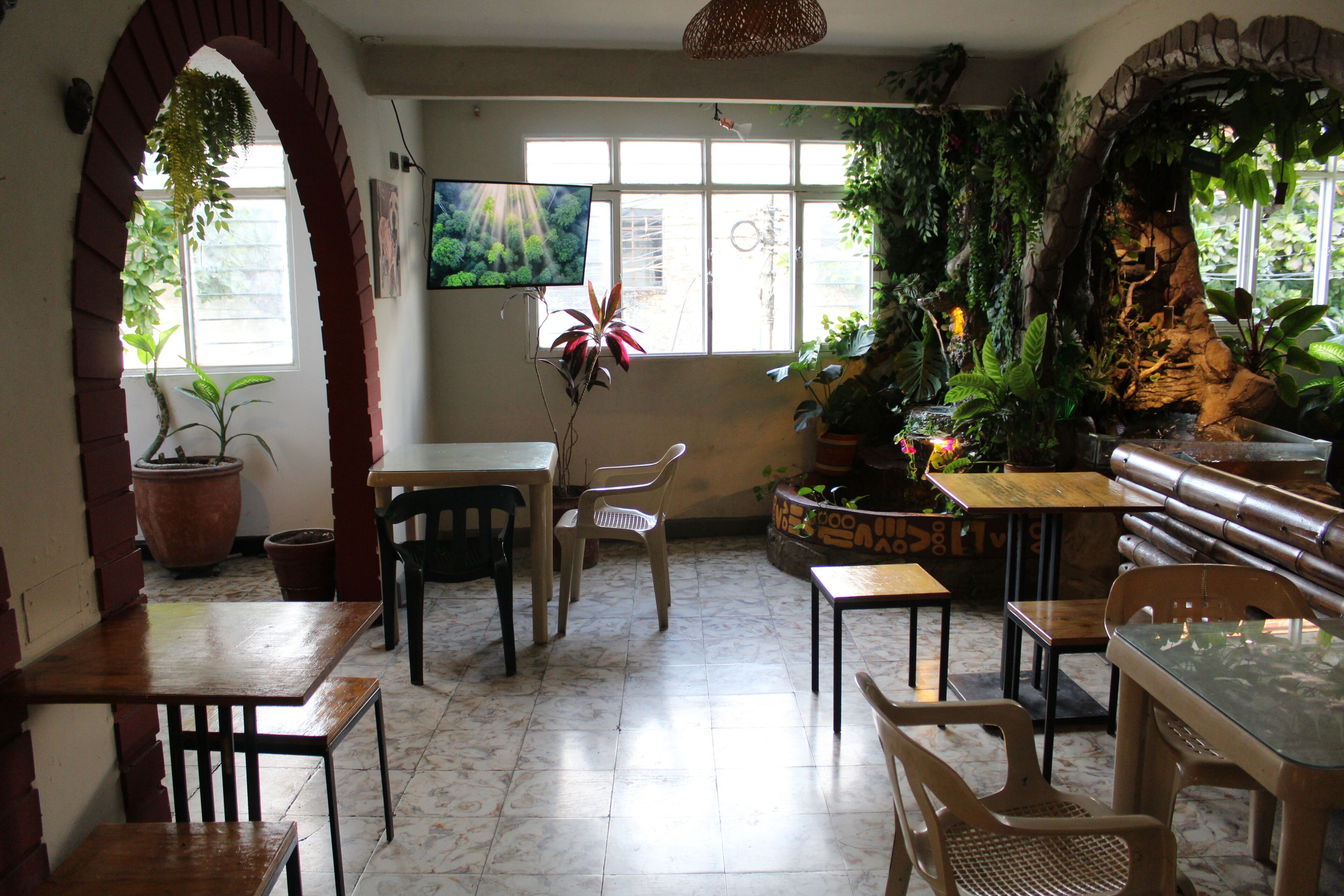 A restaurant interior with brick arches, tiled flooring, wooden tables and chairs, potted plants, and a television screen.