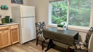 Une cuisine compacte équipée d’un réfrigérateur, de meubles en bois, d’une table à manger avec des chaises à motifs floraux et d’une fenêtre donnant sur les arbres.