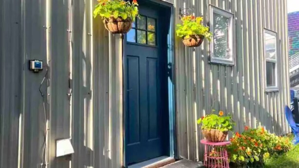 Un bâtiment en métal ondulé avec une porte bleue, deux jardinières suspendues et un petit tabouret rose sur lequel est posée une plante.