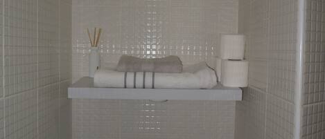Un baño con paredes de azulejos blancos, un inodoro y un estante con artículos de aseo.