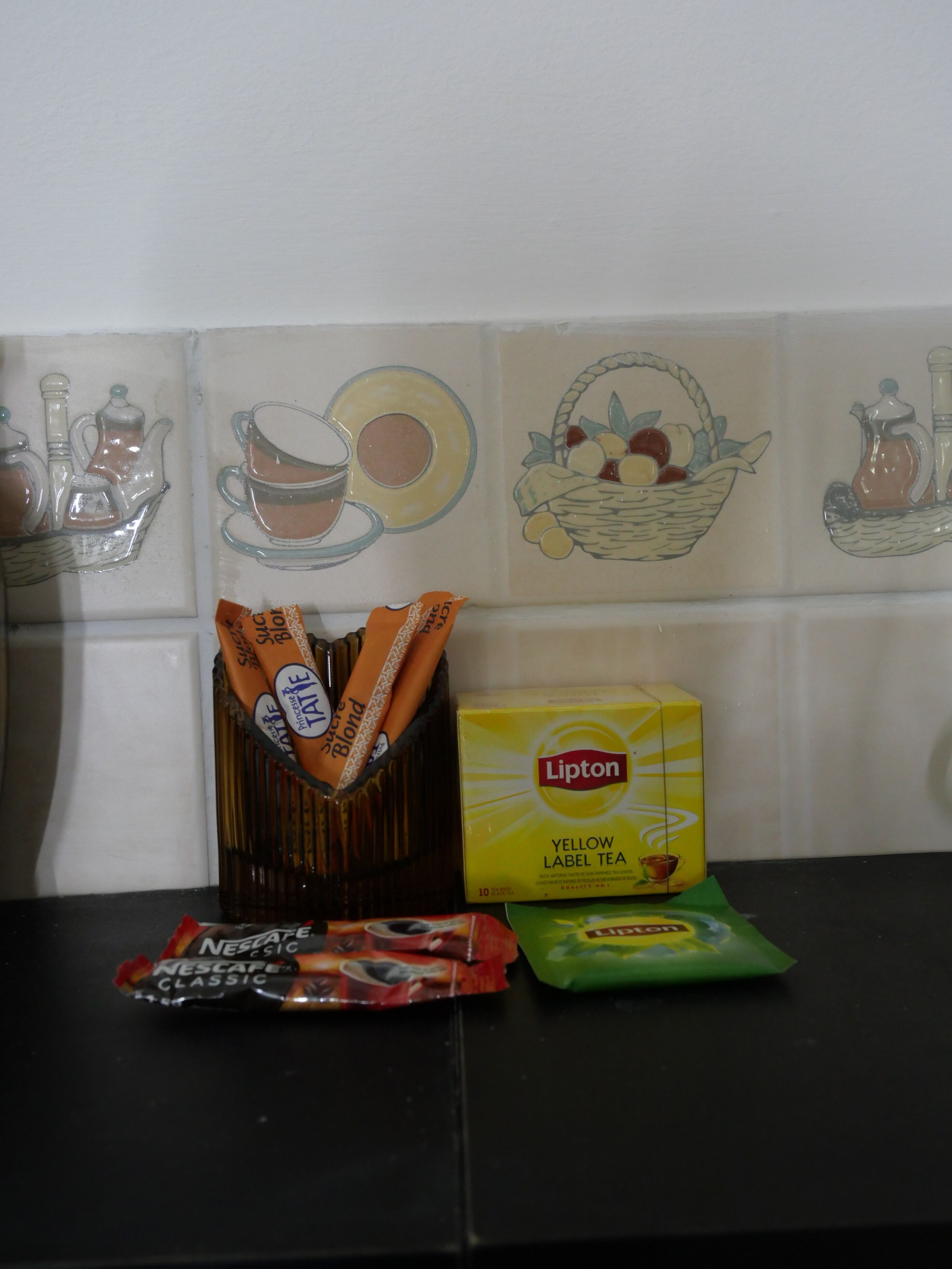 A kitchen counter with a box of Lipton Yellow Label Tea, a packet of Nescafe Classic coffee, and a packet of Lipton tea.