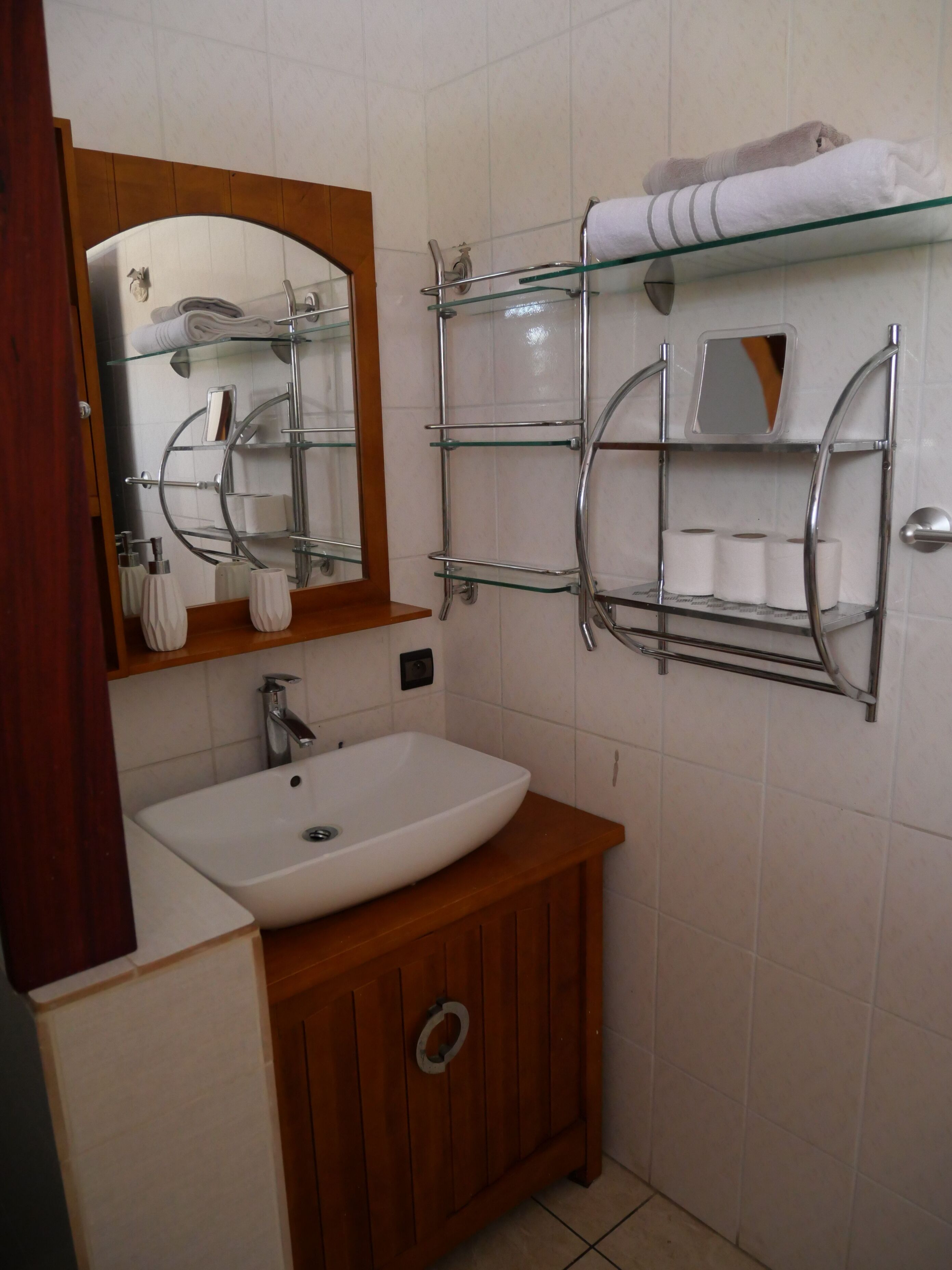 A bathroom with a white sink, wooden cabinet, and a glass shelf with towels and toiletries.