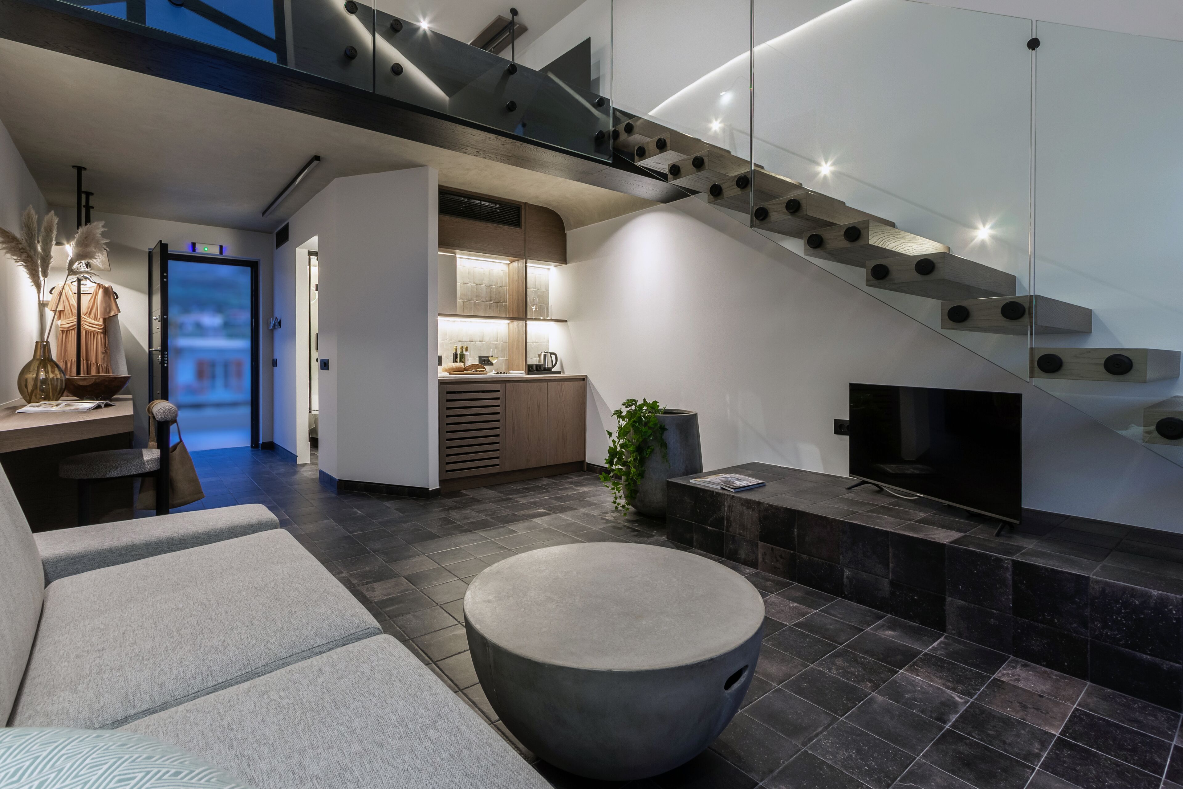 A modern living room with a grey sofa, a round ottoman, a flat-screen TV, and a staircase with a glass railing.