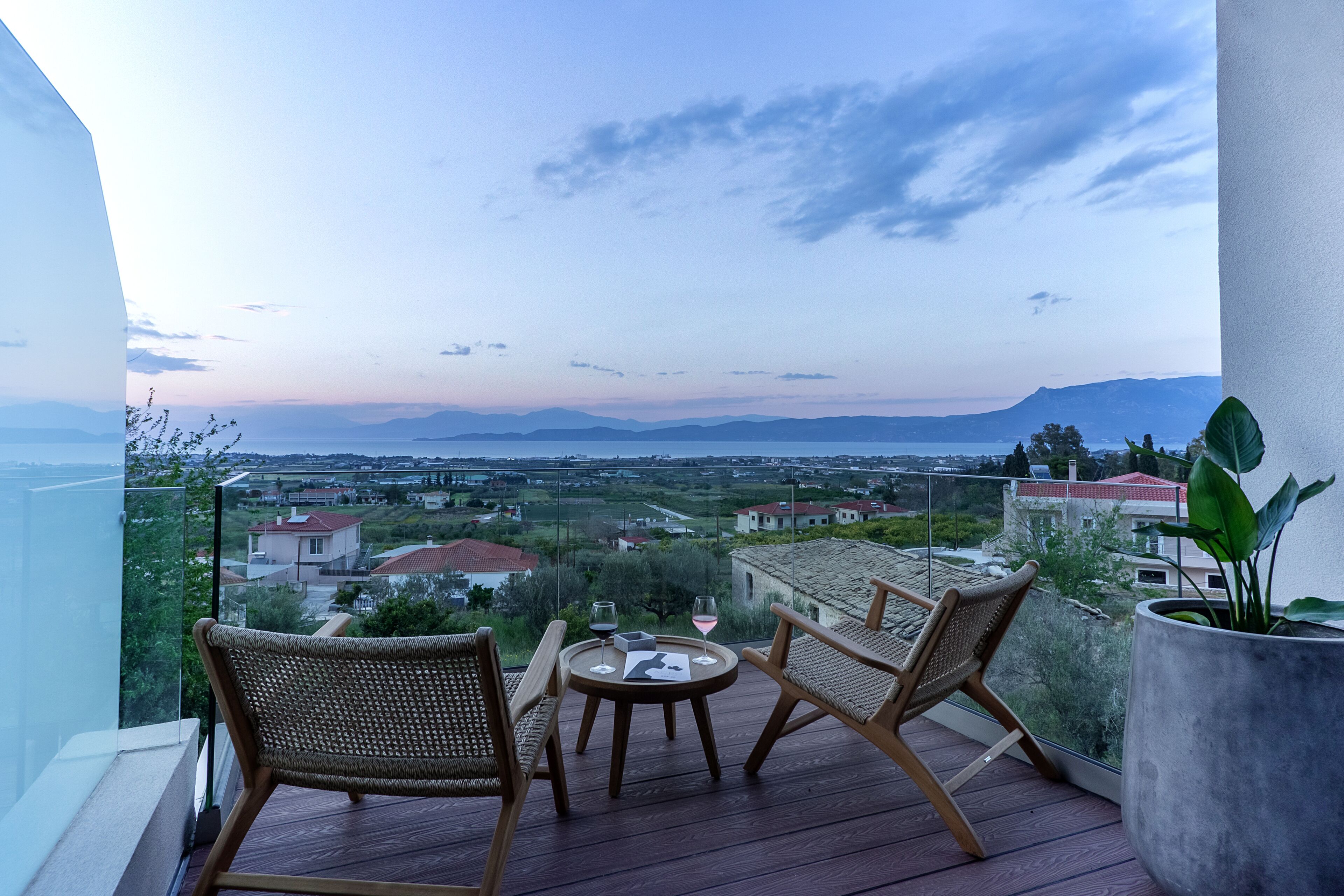 A balcony with wicker chairs and a table, overlooking a scenic view of a town and mountains.