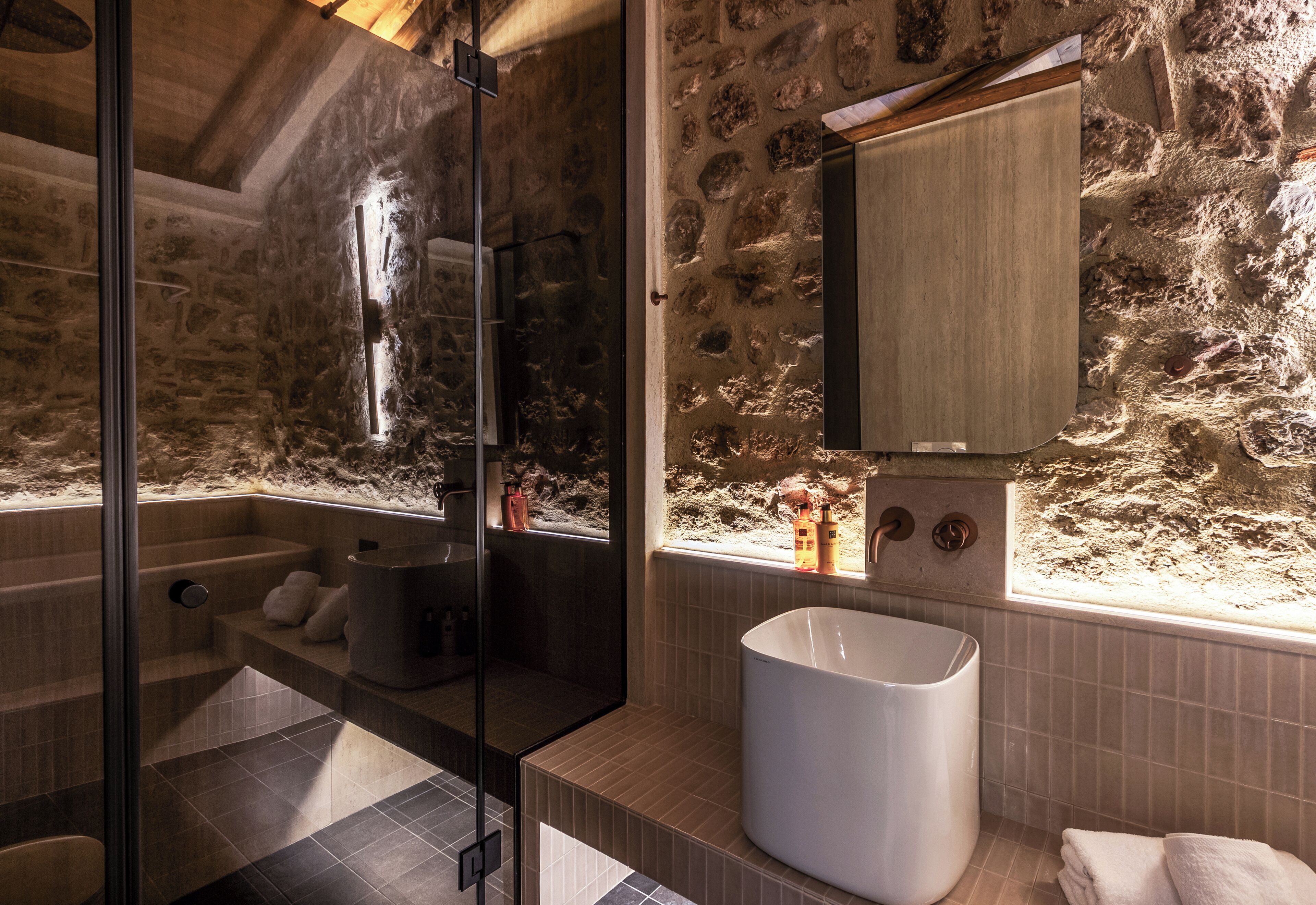 A bathroom with a stone wall, a large bathtub, a glass shower enclosure, and a sink with a mirror.