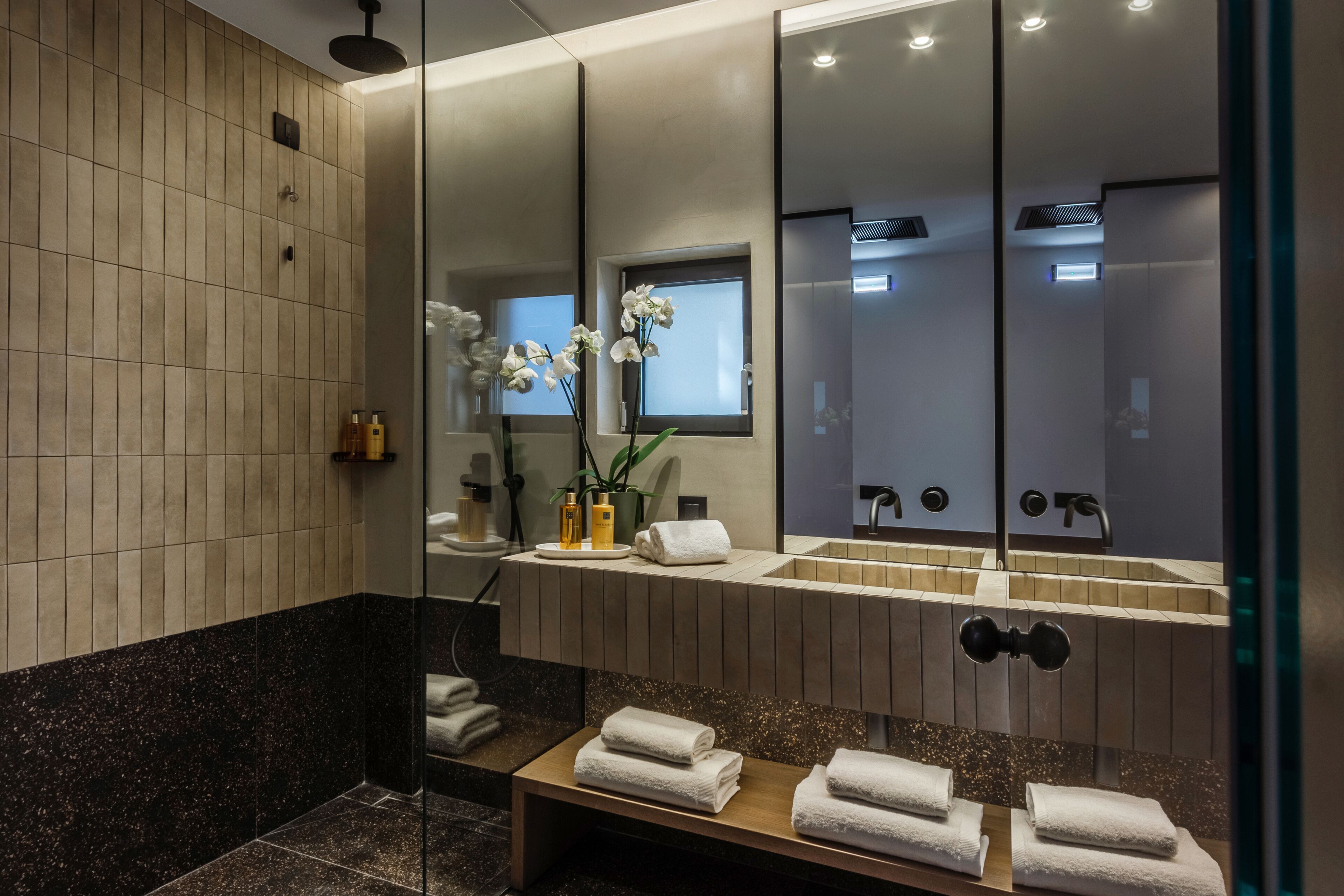 A modern bathroom with a large mirror, two sinks, a shower area, and a bench with towels.