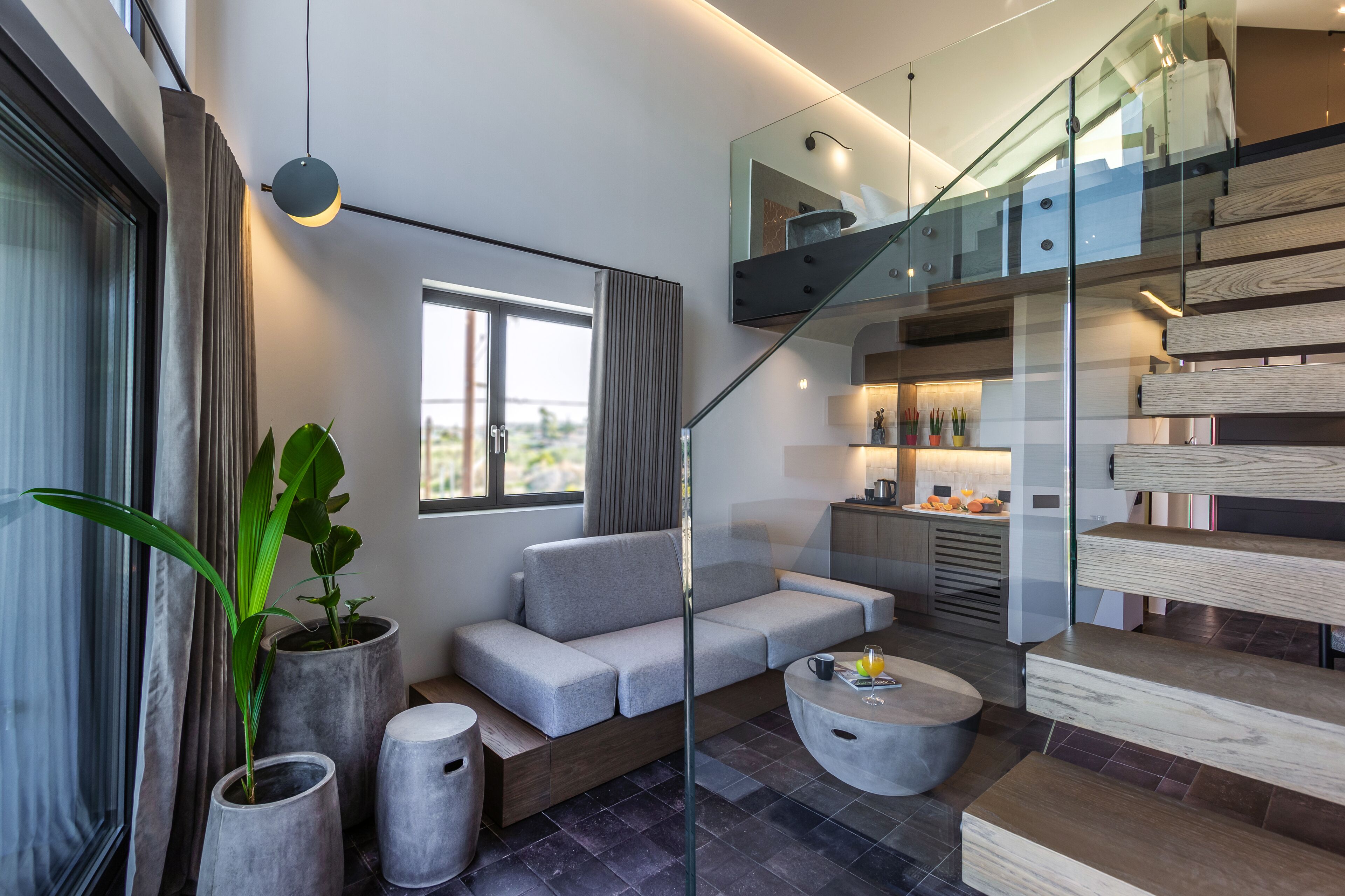 Un área de estar moderna con un sofá gris, una mesa redonda de centro y una escalera con barandillas de vidrio.