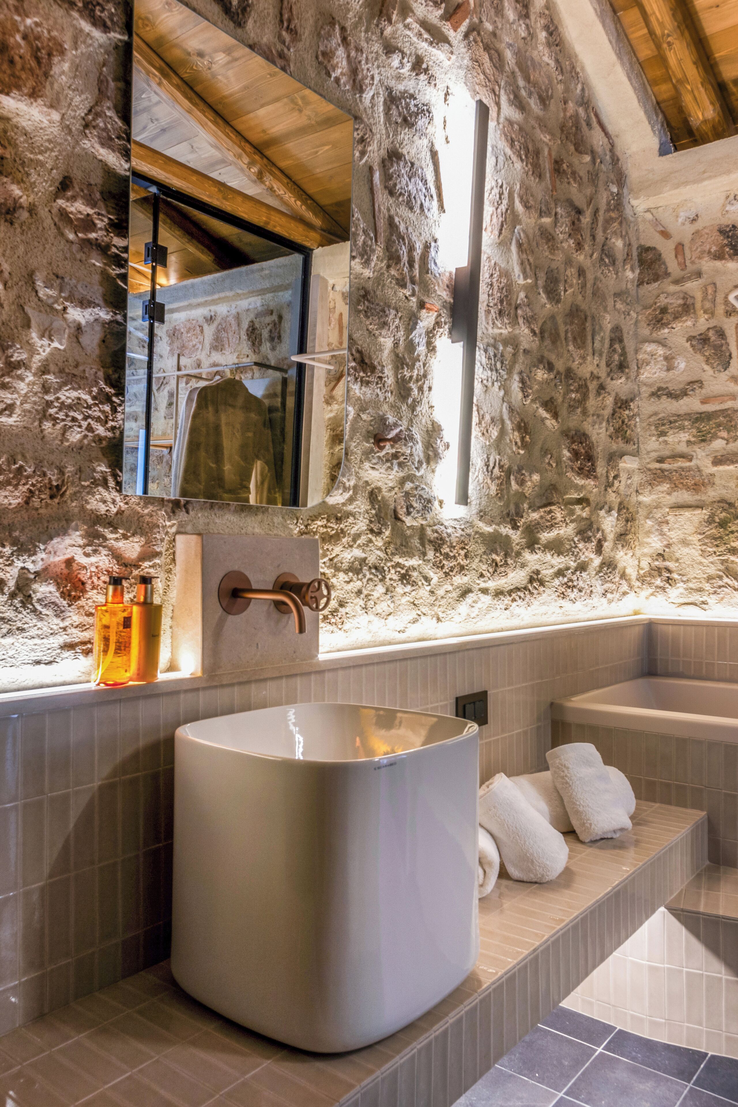 Une salle de bain avec un mur en pierre, une grande baignoire et un miroir dans lequel se reflète une serviette.