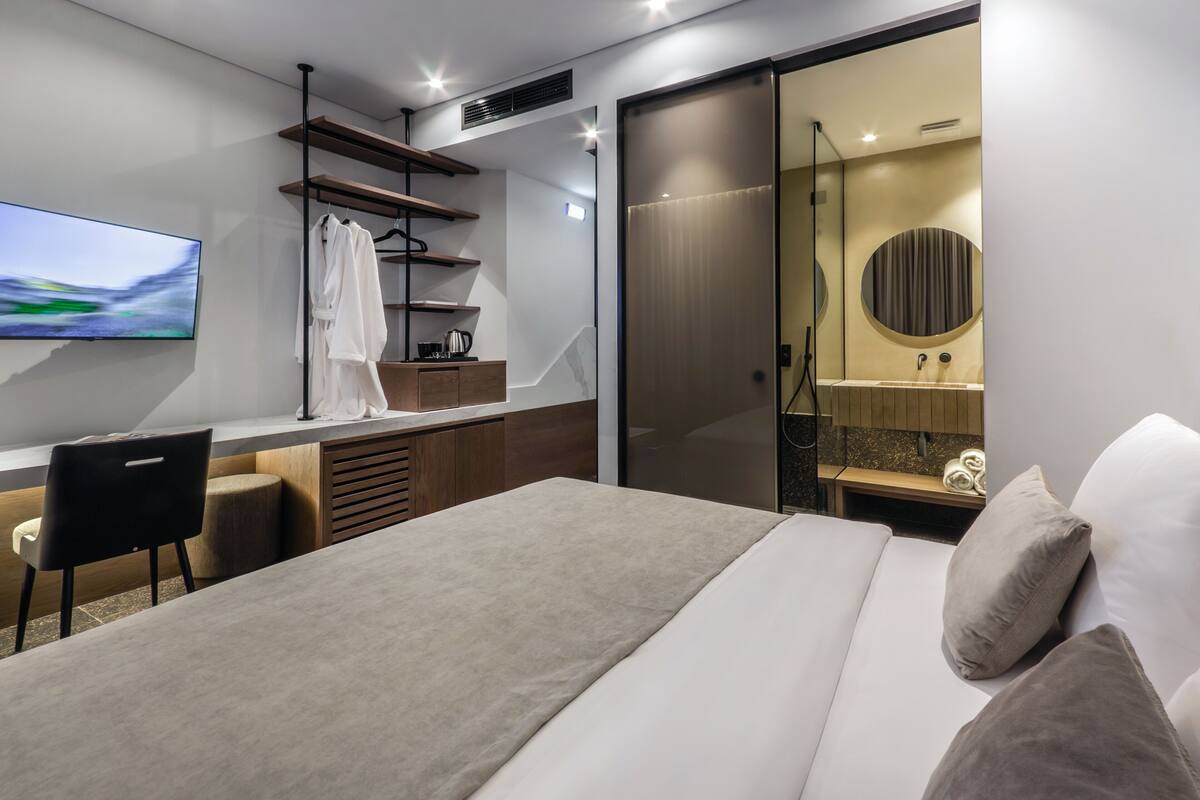 Una habitación de hotel moderna con cama, escritorio, televisor y baño con espejo y lavamanos.