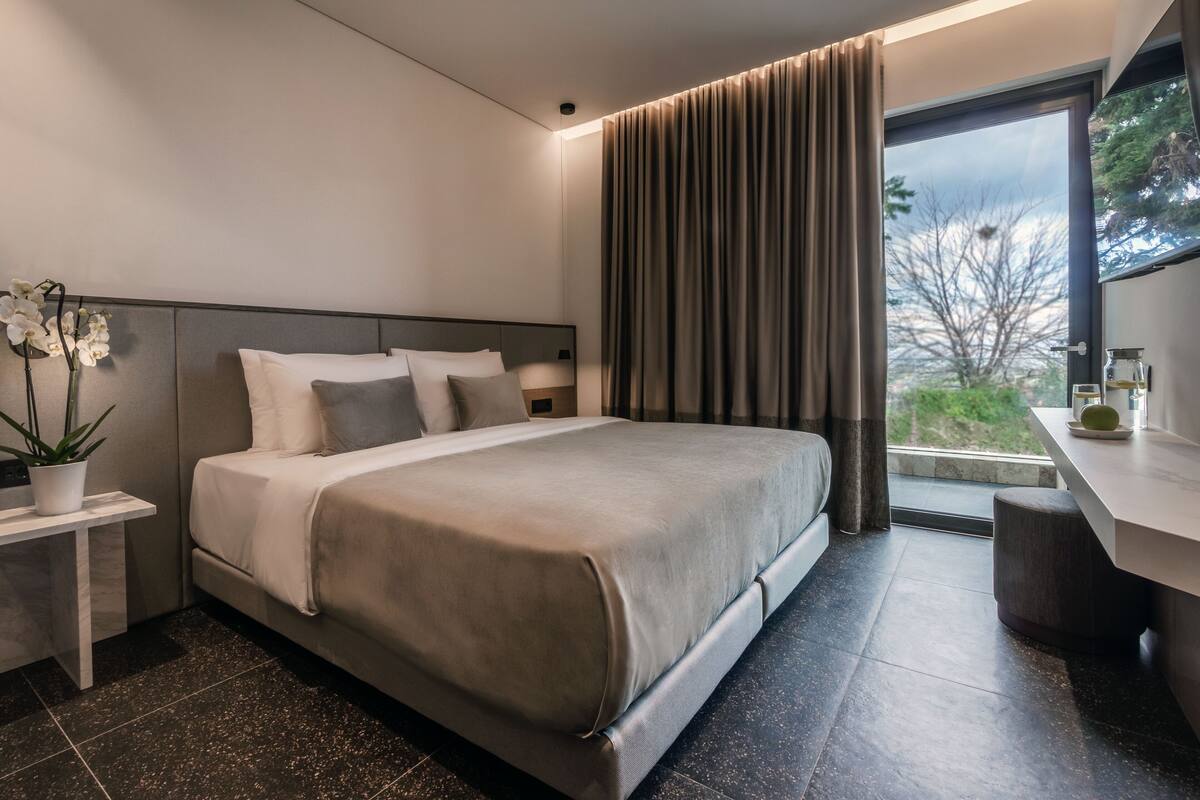 Una habitación de hotel moderna con una cama grande, una mesita de noche con una planta, un escritorio pequeño con una lámpara y una ventana con cortinas.