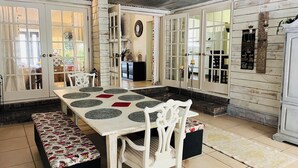 A dining area with a table set for four, surrounded by white chairs. There is a bench with a patterned tablecloth. The room has a high ceiling with a fan, and the walls are adorned with decorative items and a clock. - The Retreat - Entire Residential Home! (Daphne)