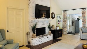 A living room with a white brick fireplace, a mounted TV, and a grey armchair. - The Retreat - Entire Residential Home! (Daphne)