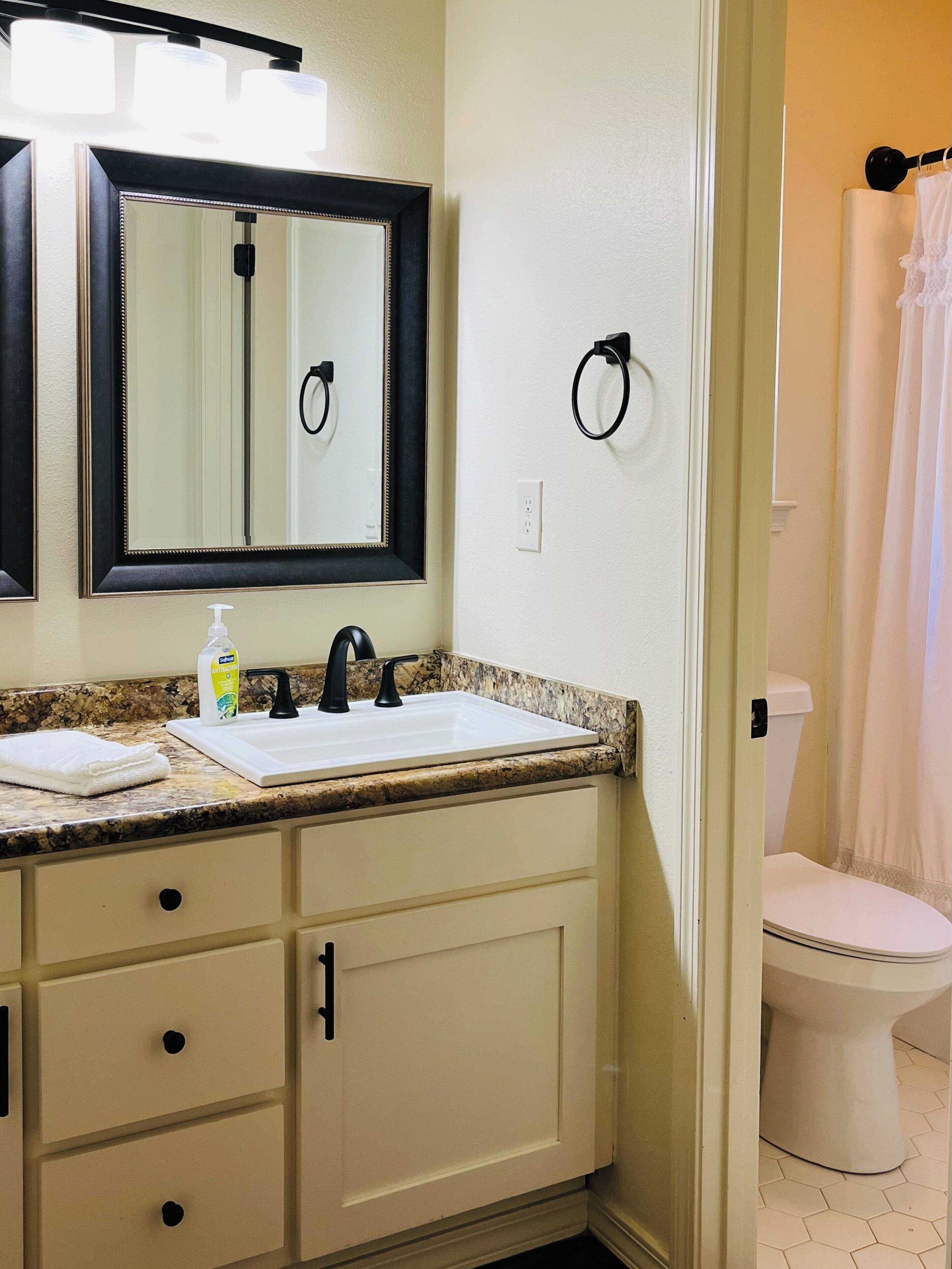 Une salle de bain avec un lavabo blanc, un robinet noir et un miroir avec un cadre noir. Il y a une serviette sur le comptoir et un flacon de savon liquide. Les murs sont peints dans une couleur claire, et on aperçoit des toilettes au fond.