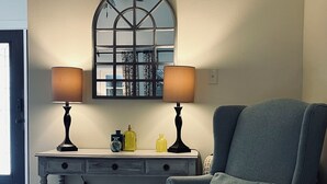 A room with a blue armchair, a desk with two lamps, a mirror, and a window with a view. - The Retreat - Entire Residential Home! (Daphne)