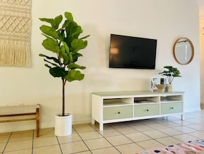 Een woonkamer met een witte bank, een potplant, een witte kast met lades, een flatscreen televisie en een ronde spiegel aan de muur.