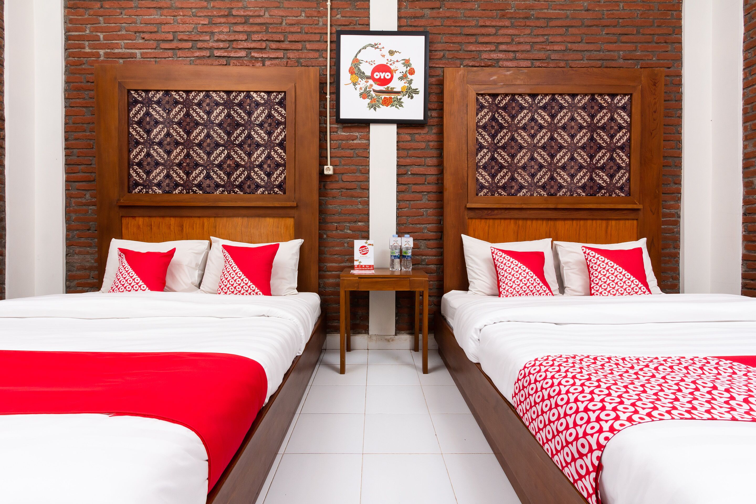 A hotel room with two beds, each with red and white bedding, a small table with a bottle and a box, and a framed picture on the wall.
